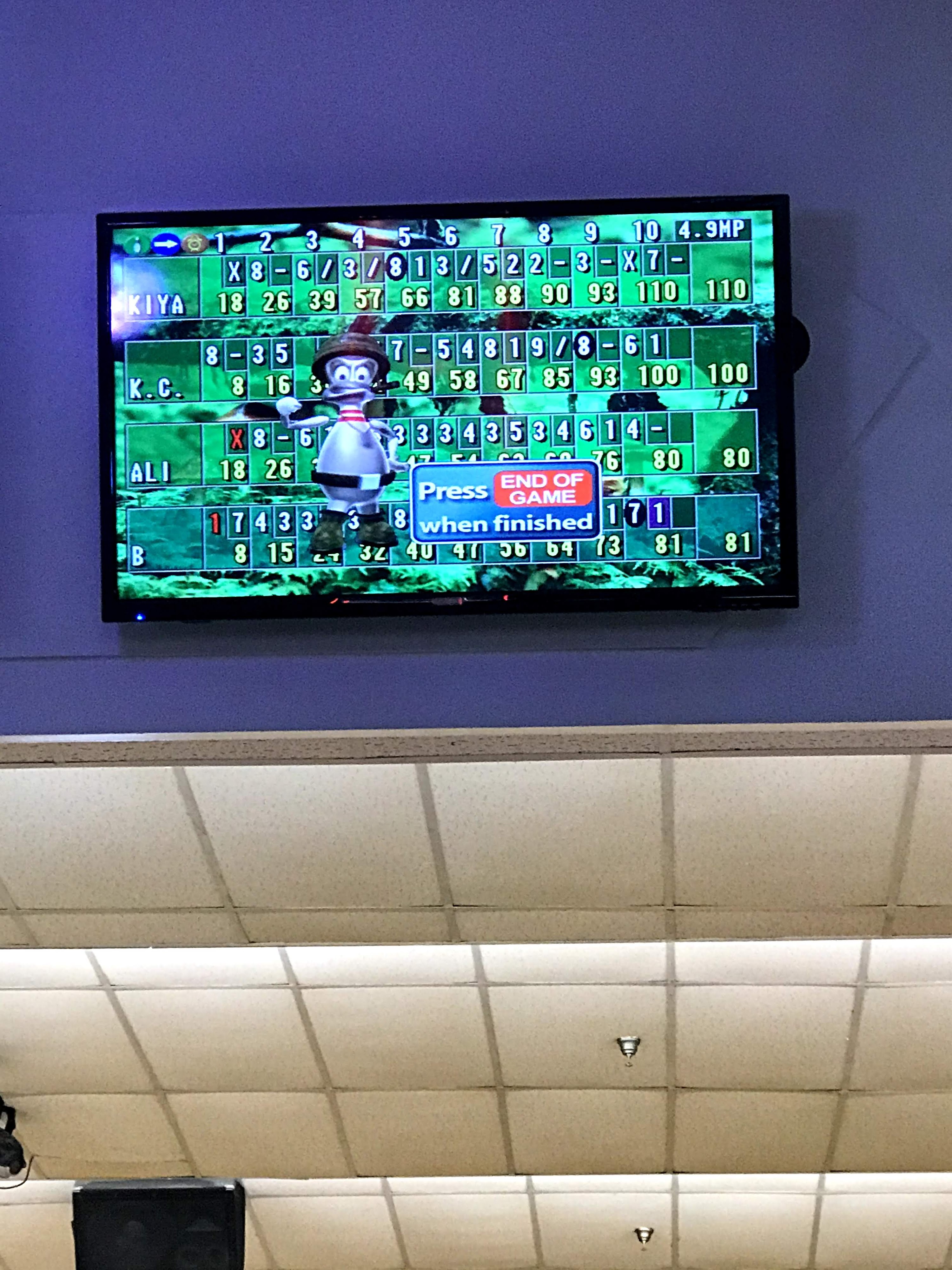 A digital bowling score monitor on a wall displaying game results for four players with a cartoon character.