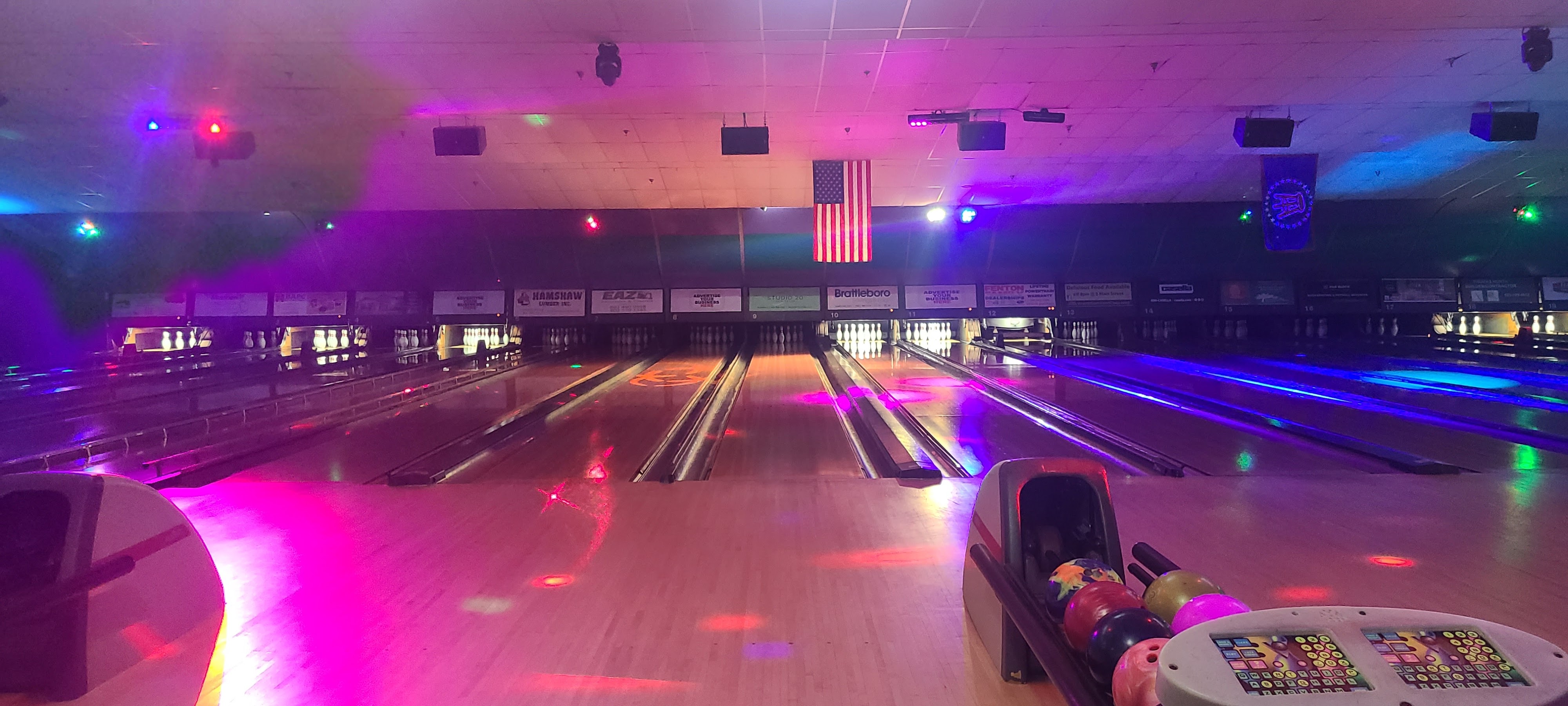 Bowling lanes at night with vibrant pink and blue neon lighting and an American flag hanging above the pins.