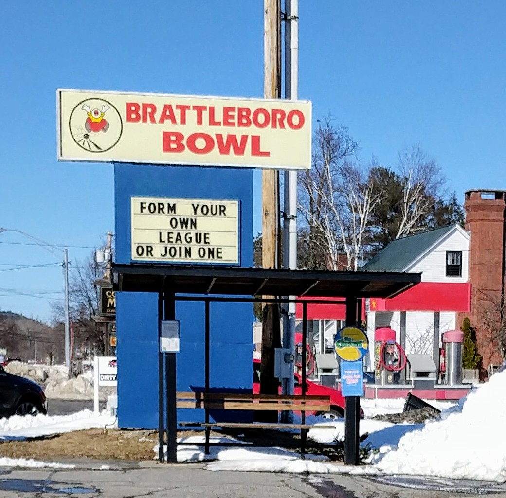 A roadside sign for Brattleboro Bowl with a marquee promoting bowling leagues next to a bus shelter.