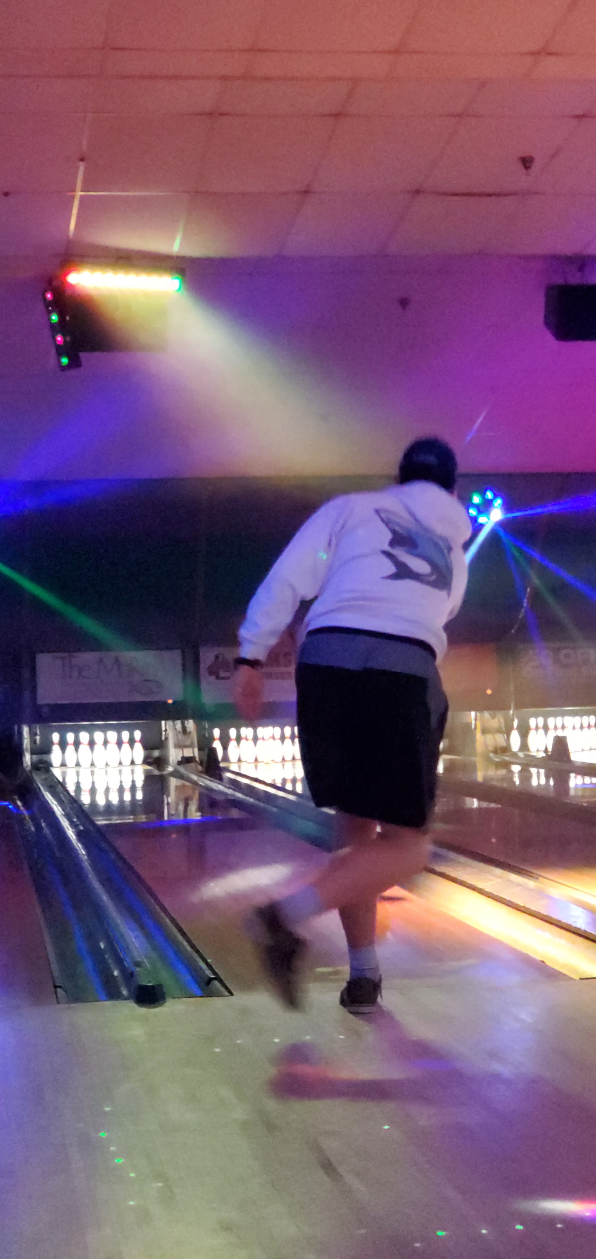 A bowler in a white hoodie takes a shot under colorful cosmic lighting on a bowling lane.