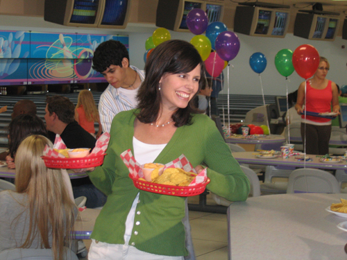 A woman smiles while holding baskets of chips and snacks at a bowling alley party decorated with colorful balloons.