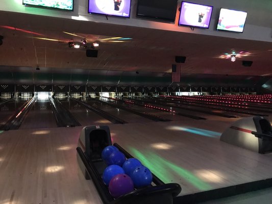 A view of bowling lanes under colorful cosmic lighting with blue and purple bowling balls in the foreground.