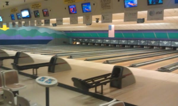 An empty bowling alley with wooden lanes, ball returns, and overhead scoring monitors under bright indoor lighting.