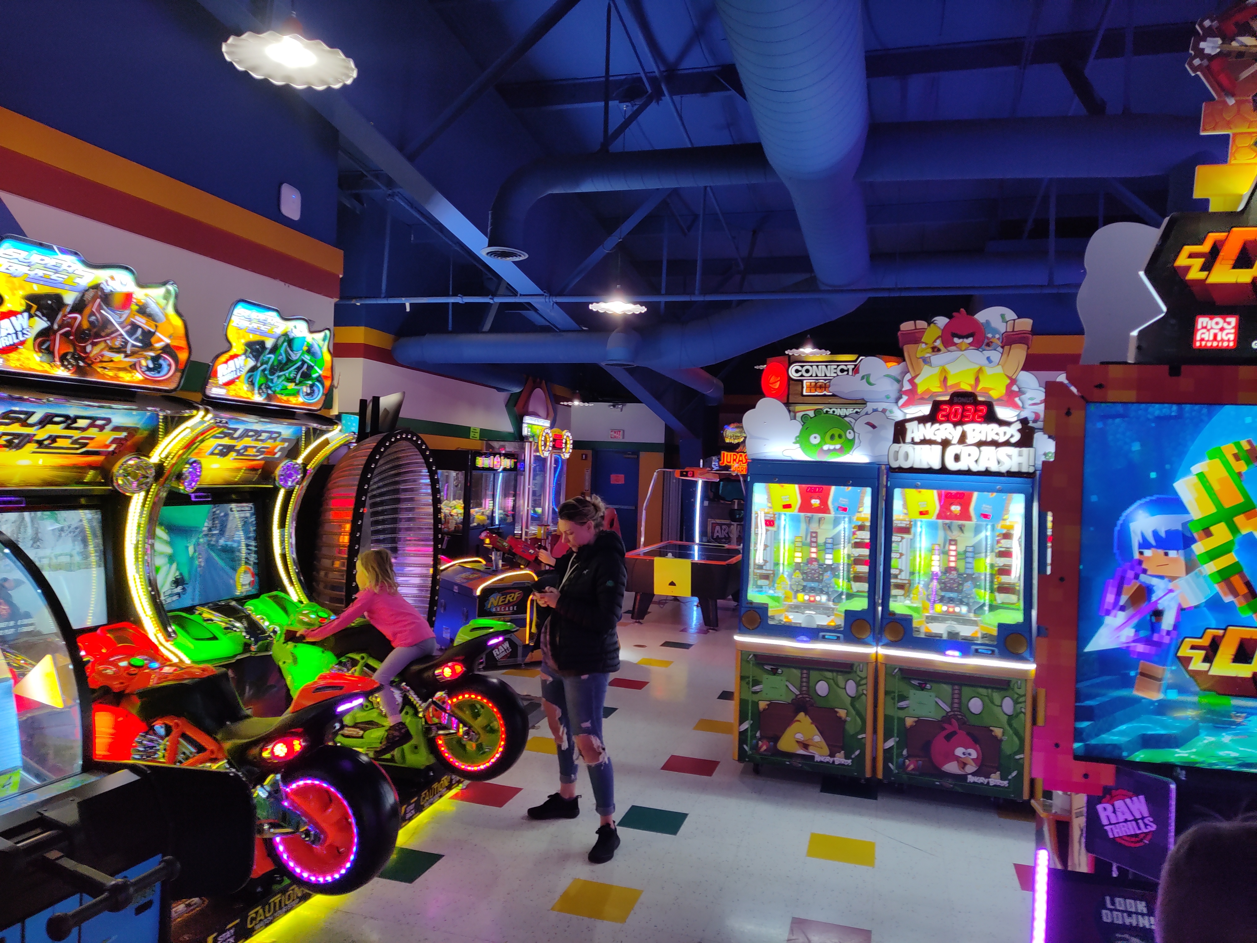 A child plays a motorcycle racing simulator in a brightly lit arcade with various modern video games and prize machines.