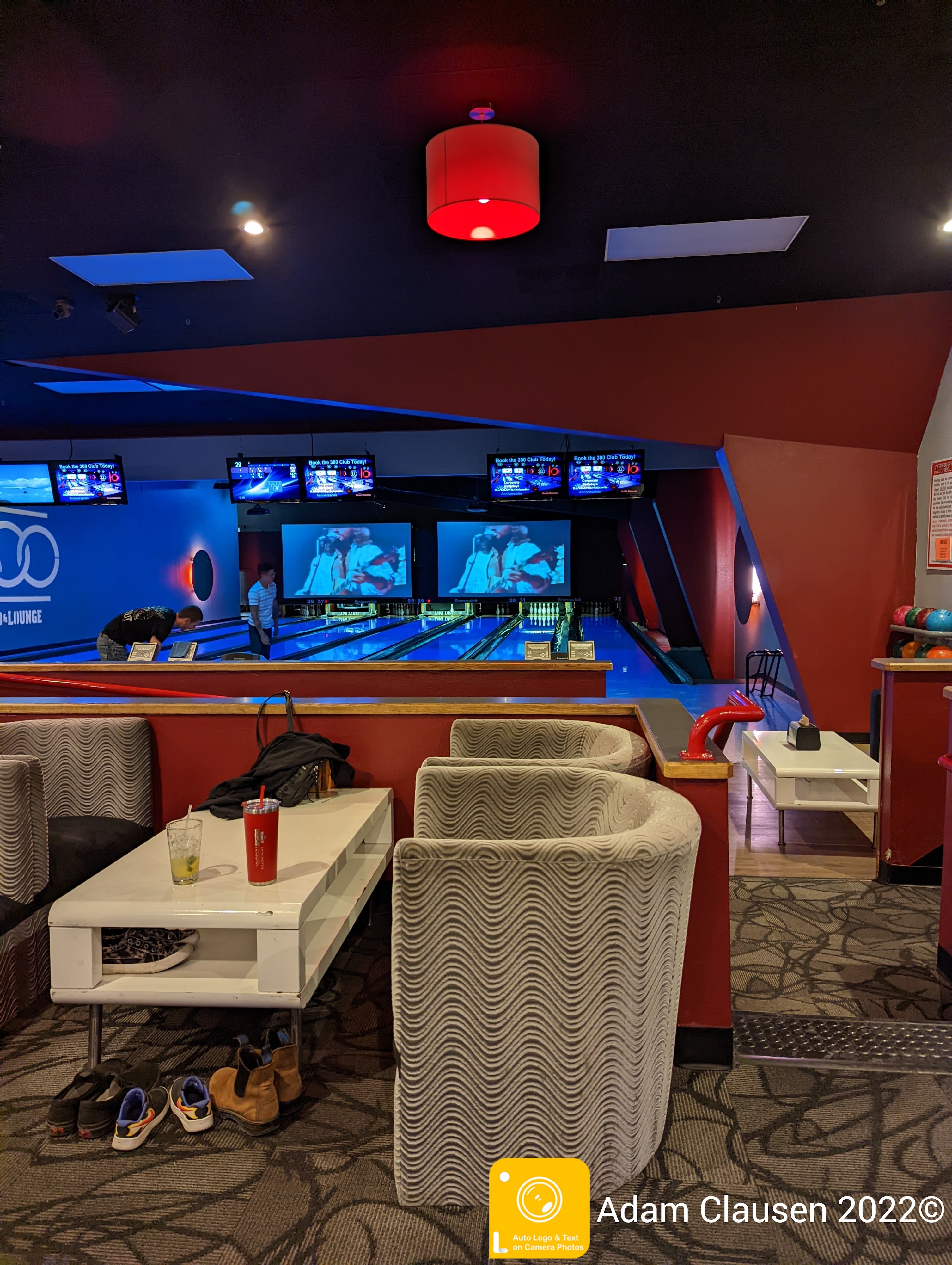 A modern bowling lounge with gray armchairs and white tables overlooking blue-lit bowling lanes and large video screens.