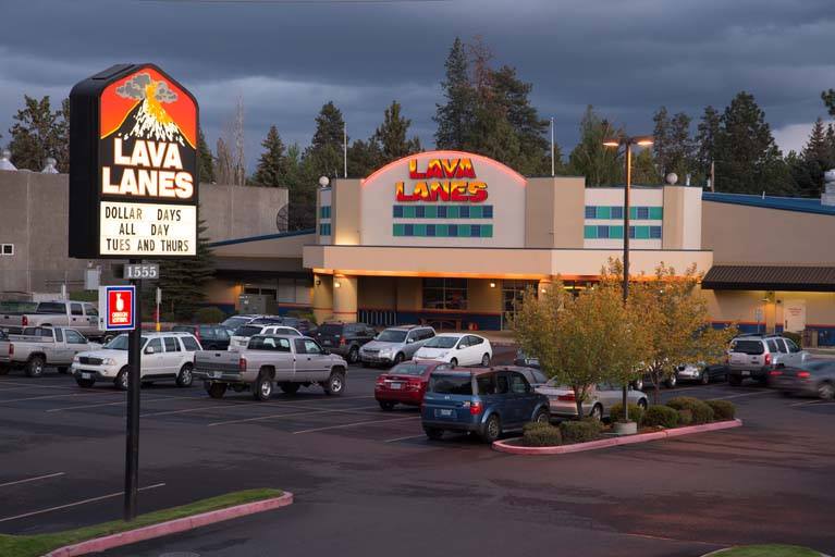 Exterior view of Lava Lanes Bend at dusk, showing the illuminated building facade, a tall sign with the Lava Lanes logo and Dollar Days promotion, and a busy parking lot filled with cars.
