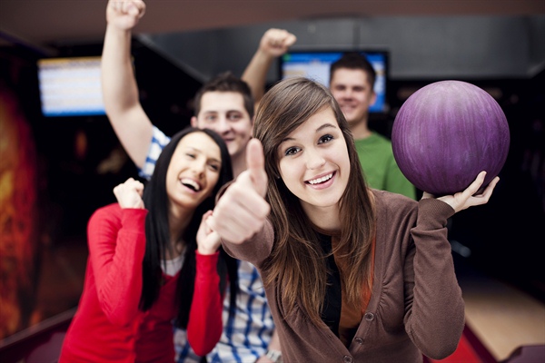 Four friends smiling and celebrating at a bowling alley as one holds a purple ball and gives a thumbs-up.