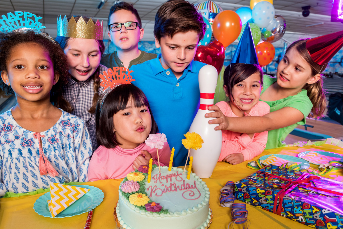 A group of smiling children in party hats gather around a birthday cake and a bowling pin at a bowling center.