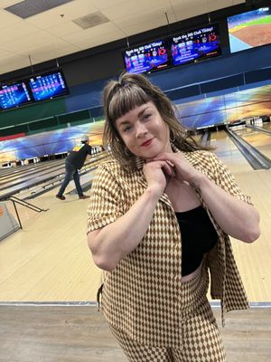 A woman in a patterned outfit poses in front of bowling lanes with a man bowling in the background.