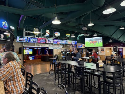 A sports bar and lounge area with high-top tables, multiple televisions, and a view into the bowling lanes.