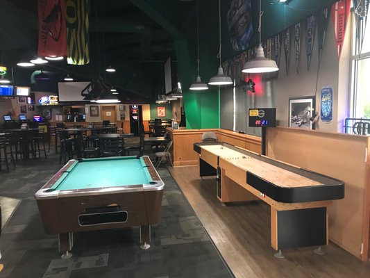 A pool table and shuffleboard table in a large recreation room with a bar and lounge in the background.
