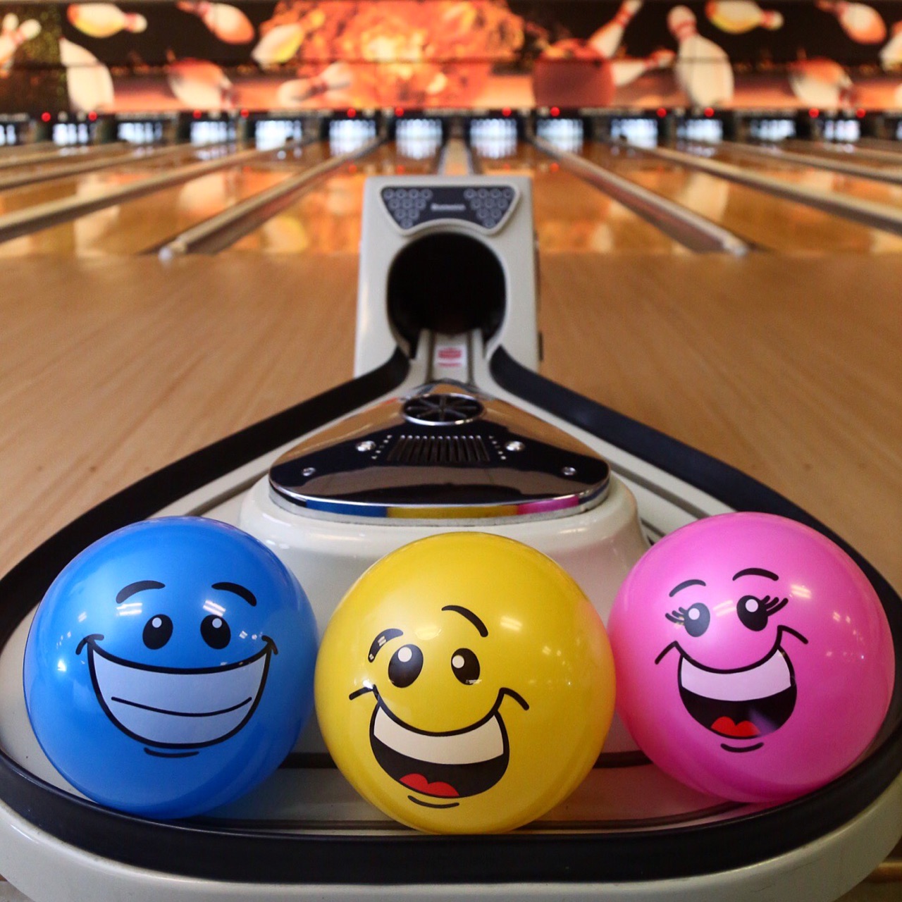Three colorful bowling balls with smiley faces sit on a ball return in front of several bowling lanes.