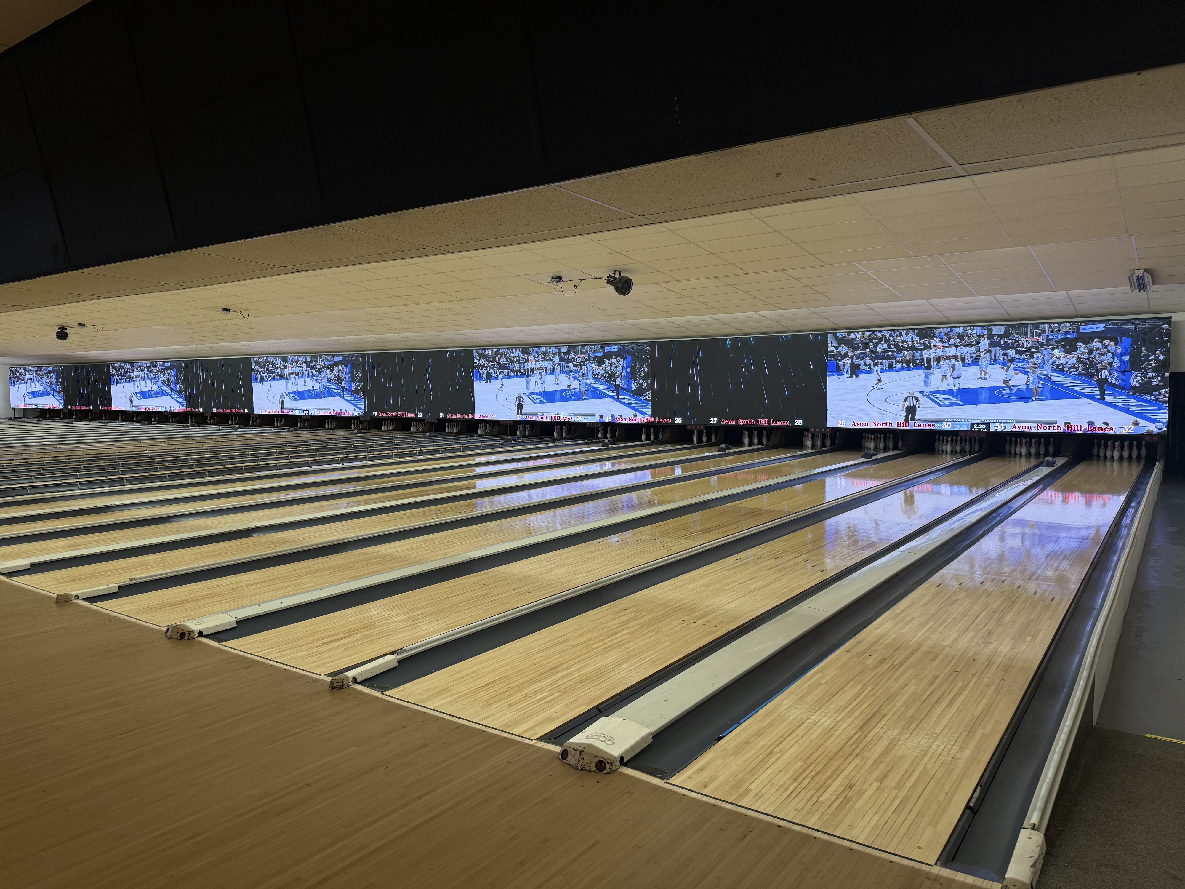 A wide view of polished bowling lanes with large video screens above the pins showing a basketball game.