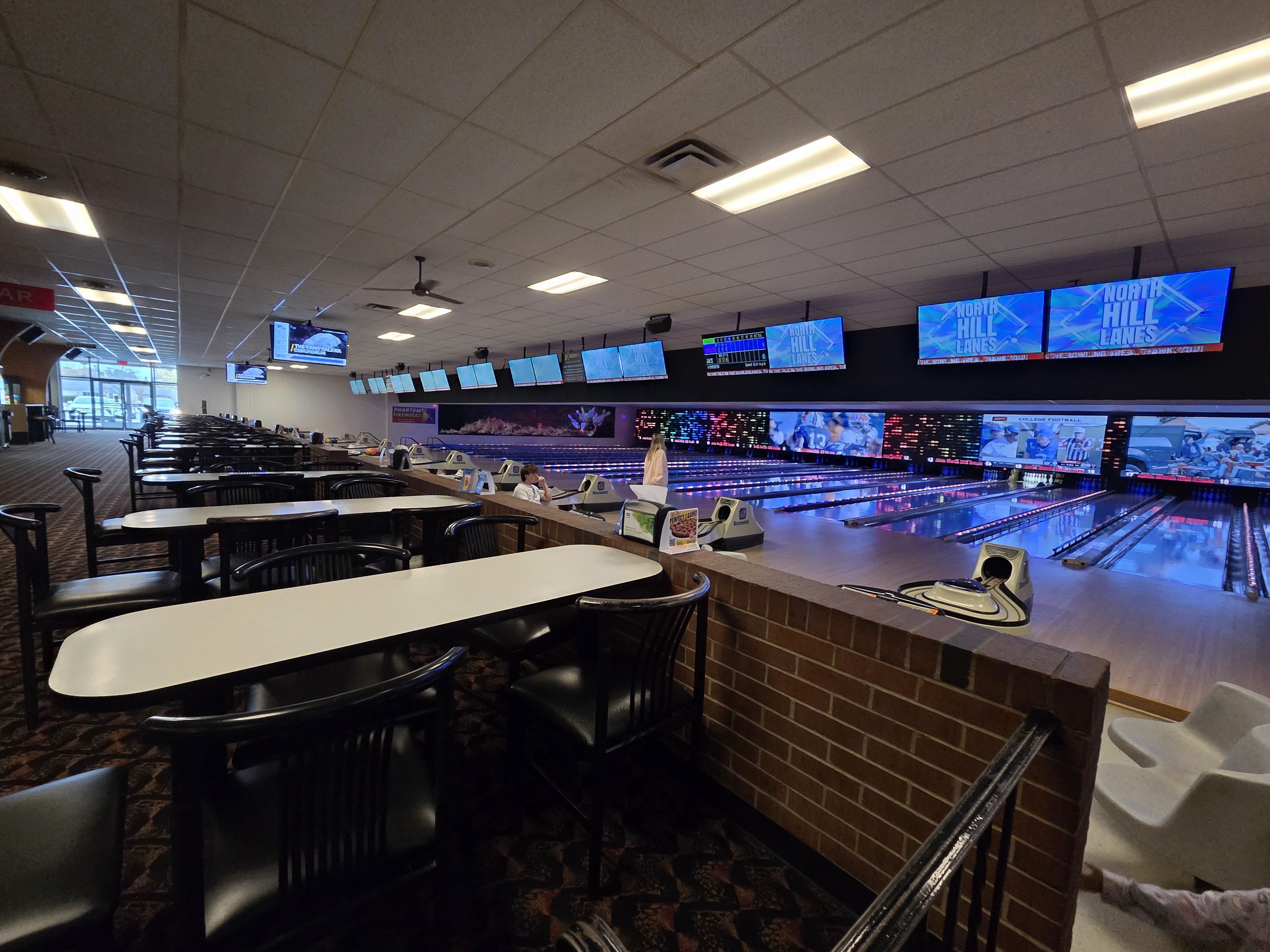A wide view of bowling lanes and a seating area with tables and chairs at a well-lit bowling center.