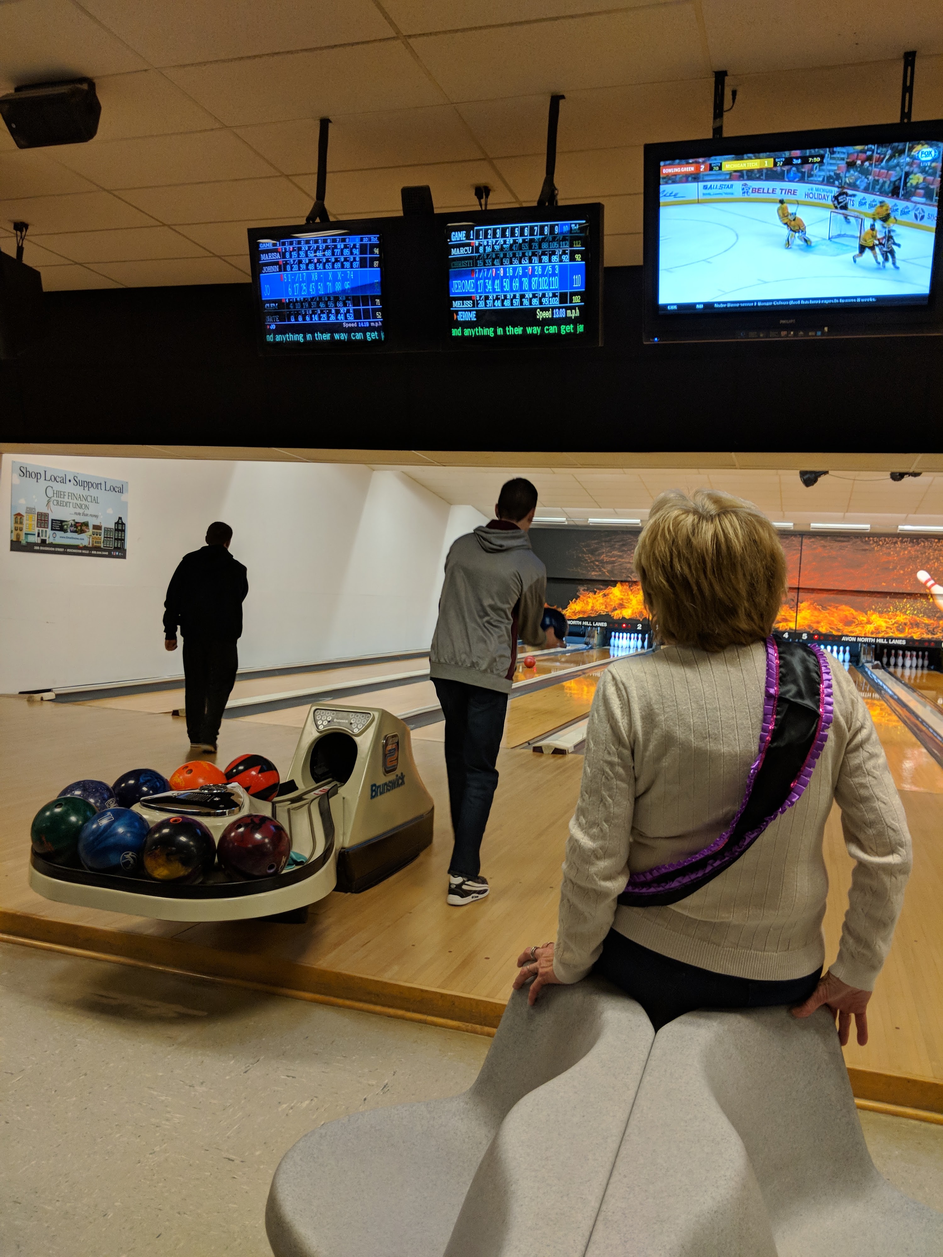 A woman in a sash watches from a bench as a man bowls on well-lit lanes with overhead scoring monitors.