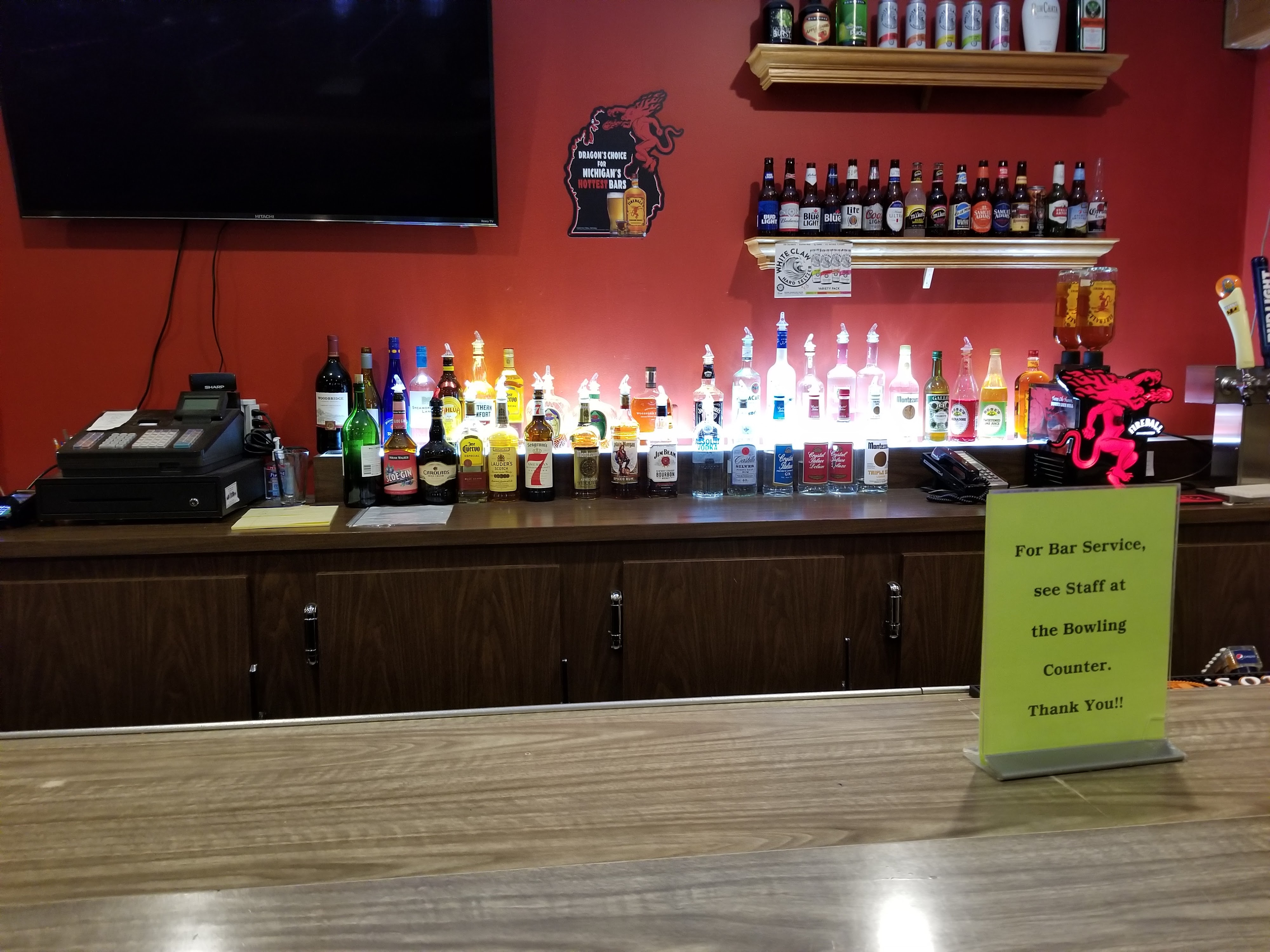 A bar counter with a red wall, illuminated liquor bottles, a television, and a green service sign.