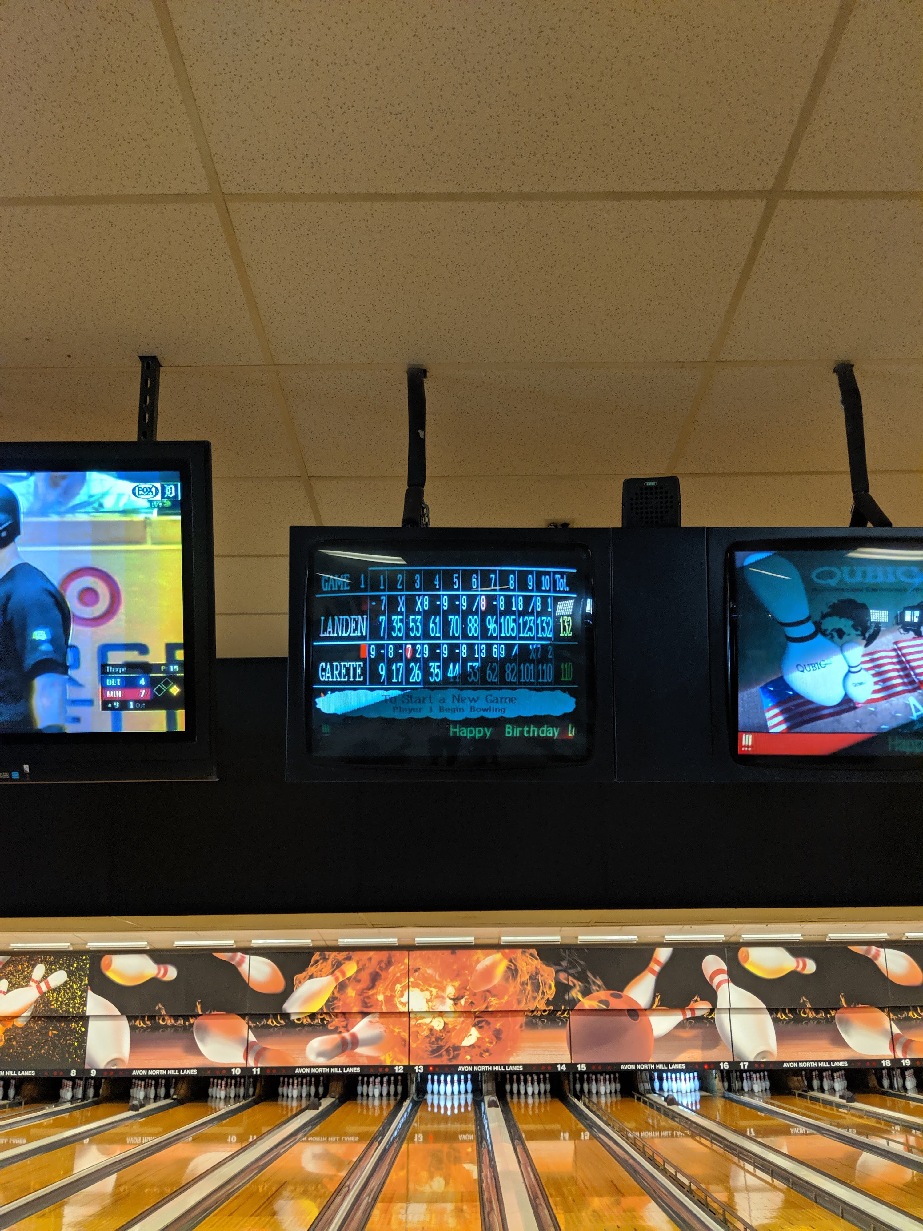 Overhead bowling monitors displaying scores and a birthday message above several lanes.