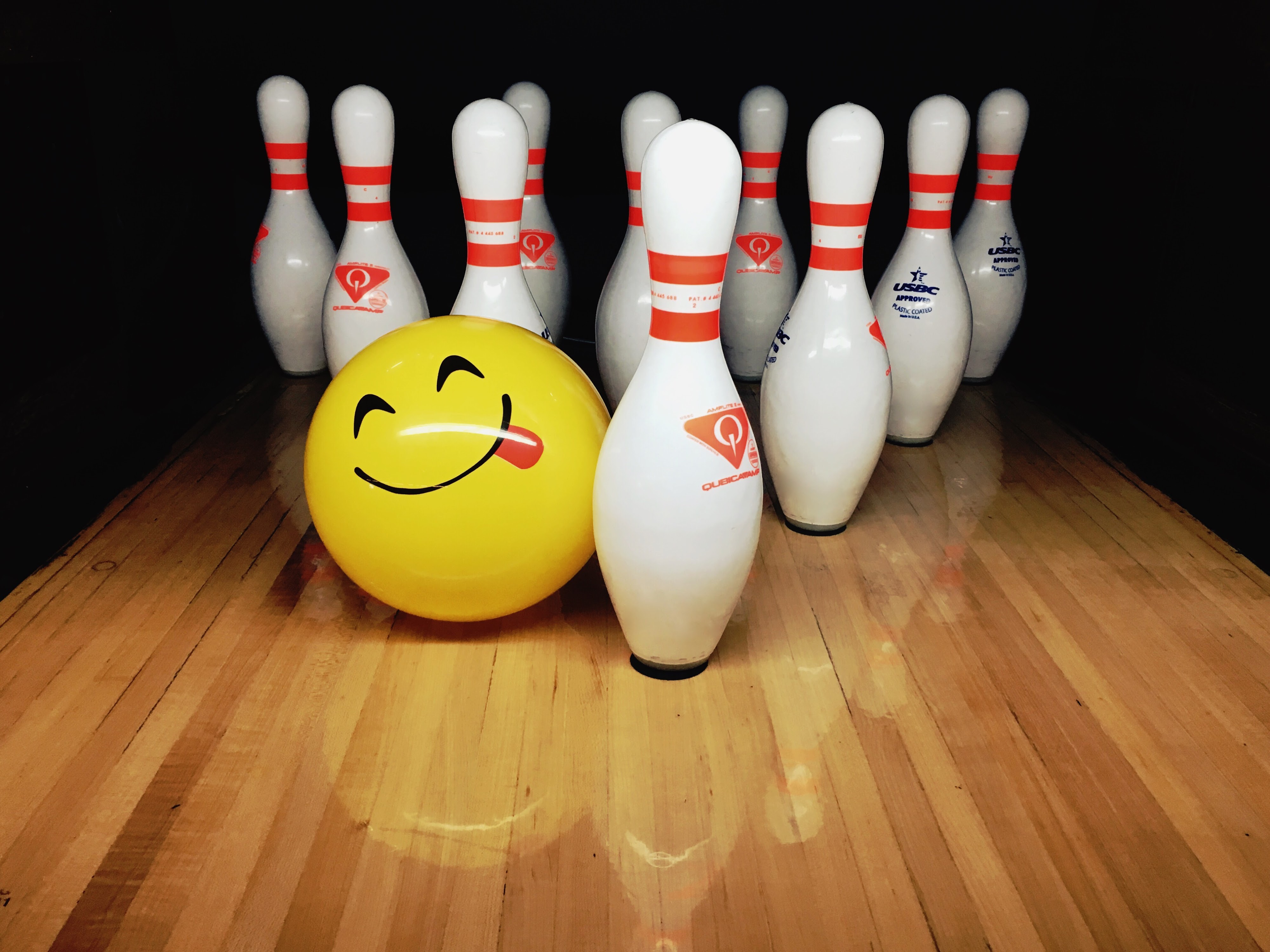 A yellow smiley face emoji bowling ball sits on a wooden lane with bowling pins.