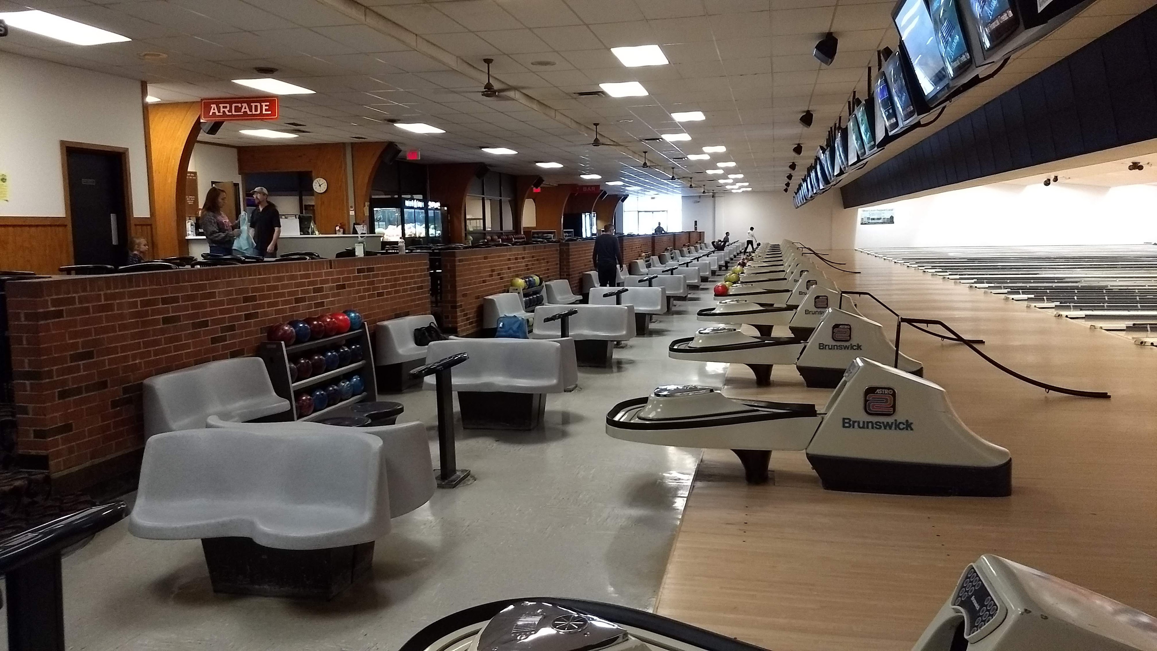 Interior view of a bowling alley featuring several lanes with grey seating, ball returns, and overhead monitors.