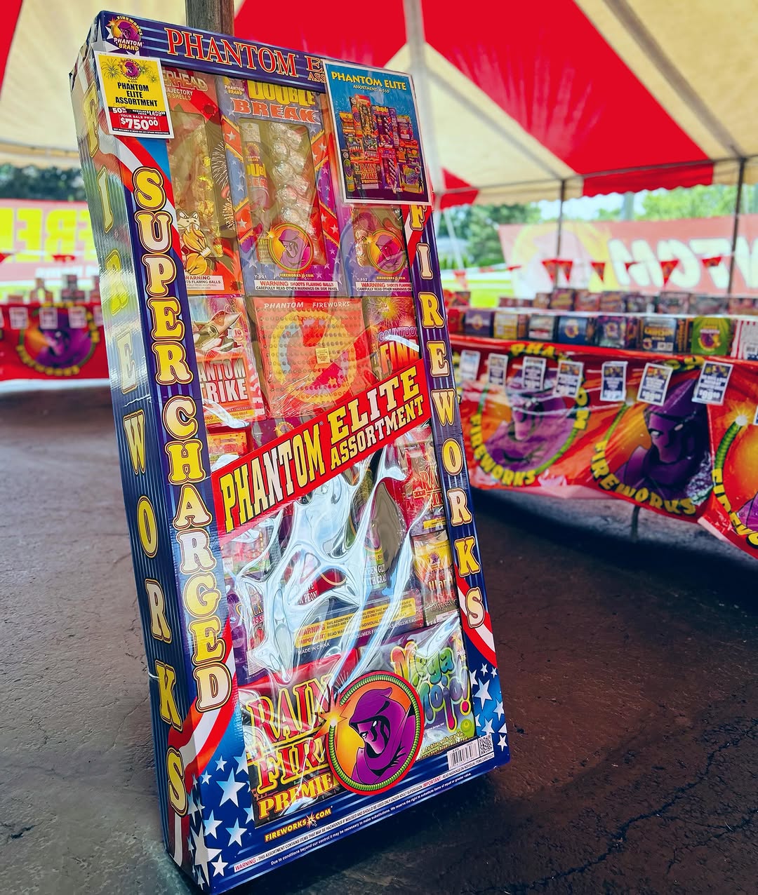 A large box of Phantom Elite Assortment fireworks stands under a red and white striped tent.