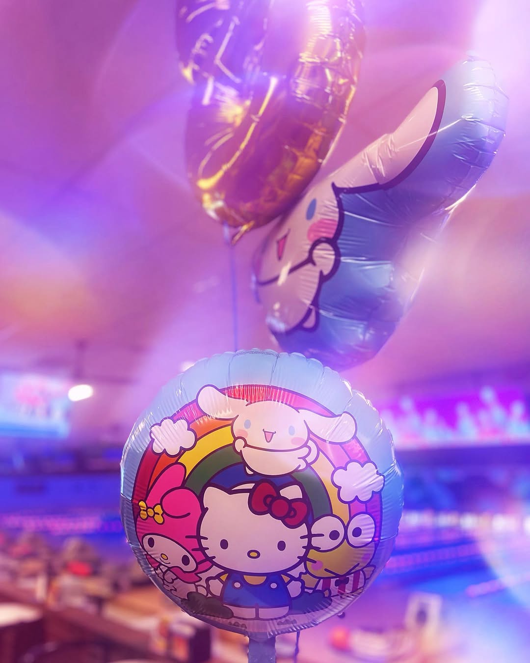 A Hello Kitty themed birthday balloon inside a bowling center with purple glow lighting and blurred lanes.