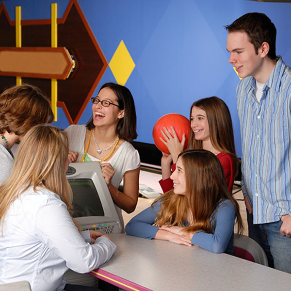 A group of five happy teenagers gathered around a bowling scoring monitor and table.