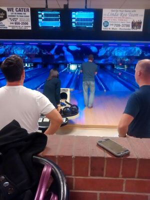 People watching a friend bowl on lanes with blue glow lighting and overhead score monitors.