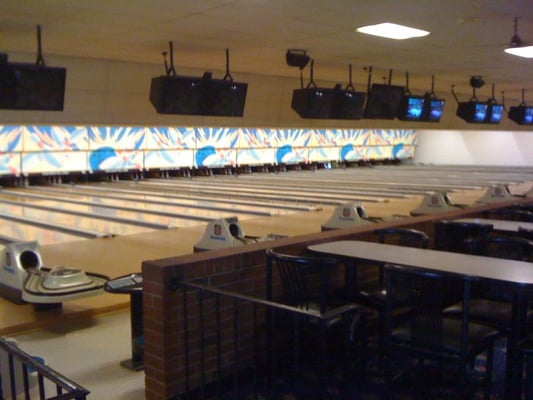 Interior view of a bowling alley with several lanes, scoring monitors, and a seating area with tables and chairs.