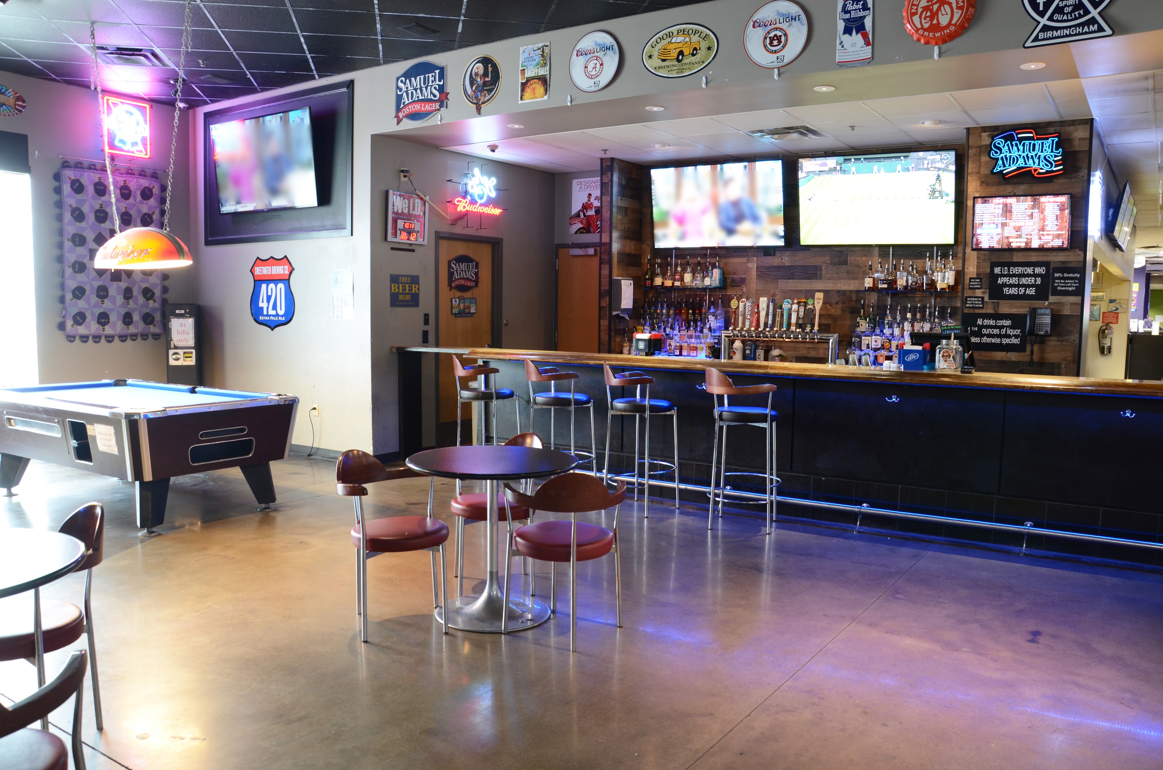A well-lit bar and lounge area with bar stools, multiple televisions, beer signage, and a blue pool table.