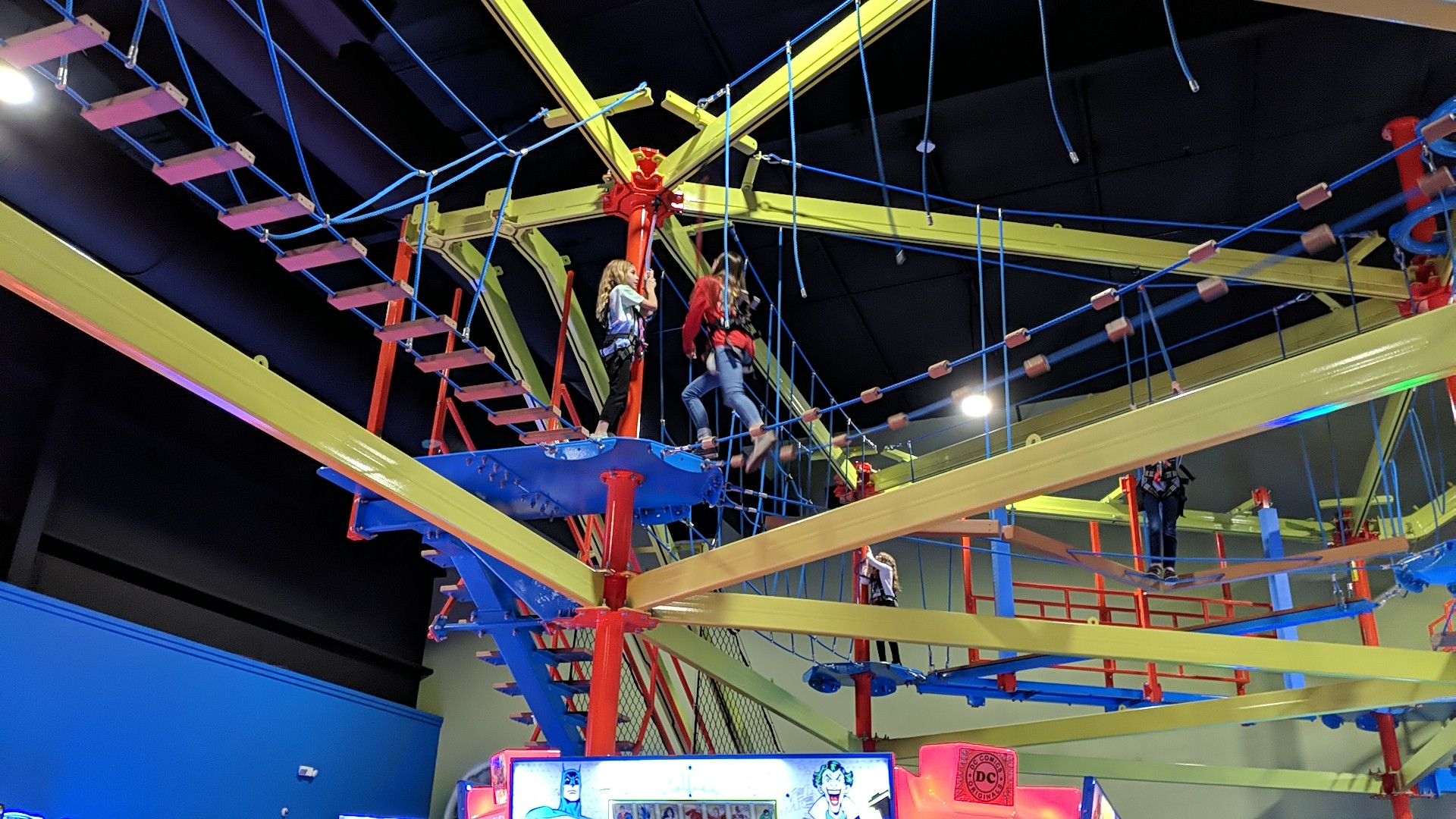 Children wearing safety harnesses navigate a colorful indoor ropes course with yellow and blue beams.
