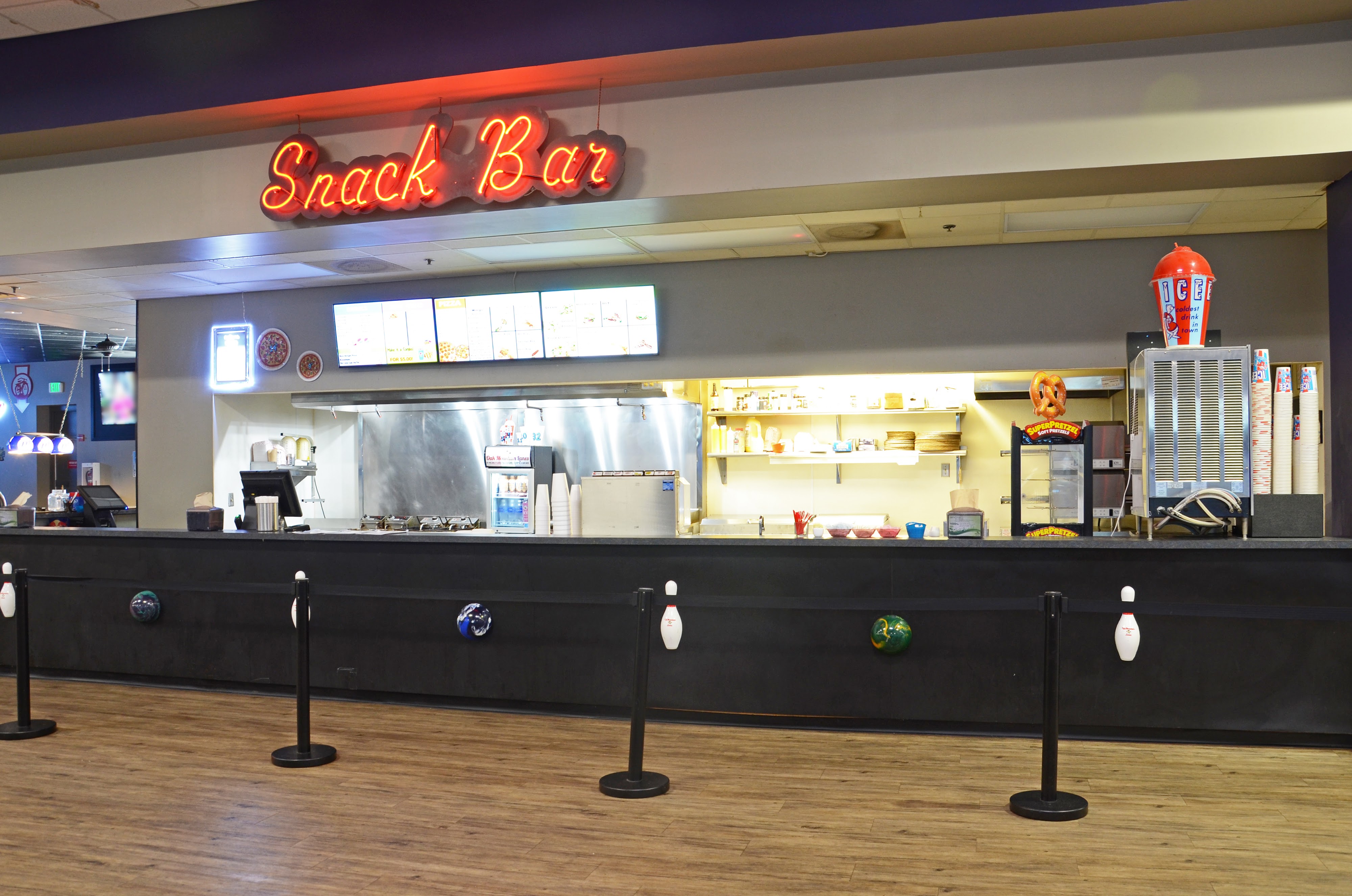 The snack bar at a bowling center featuring a red neon sign and digital menu boards above a black counter.