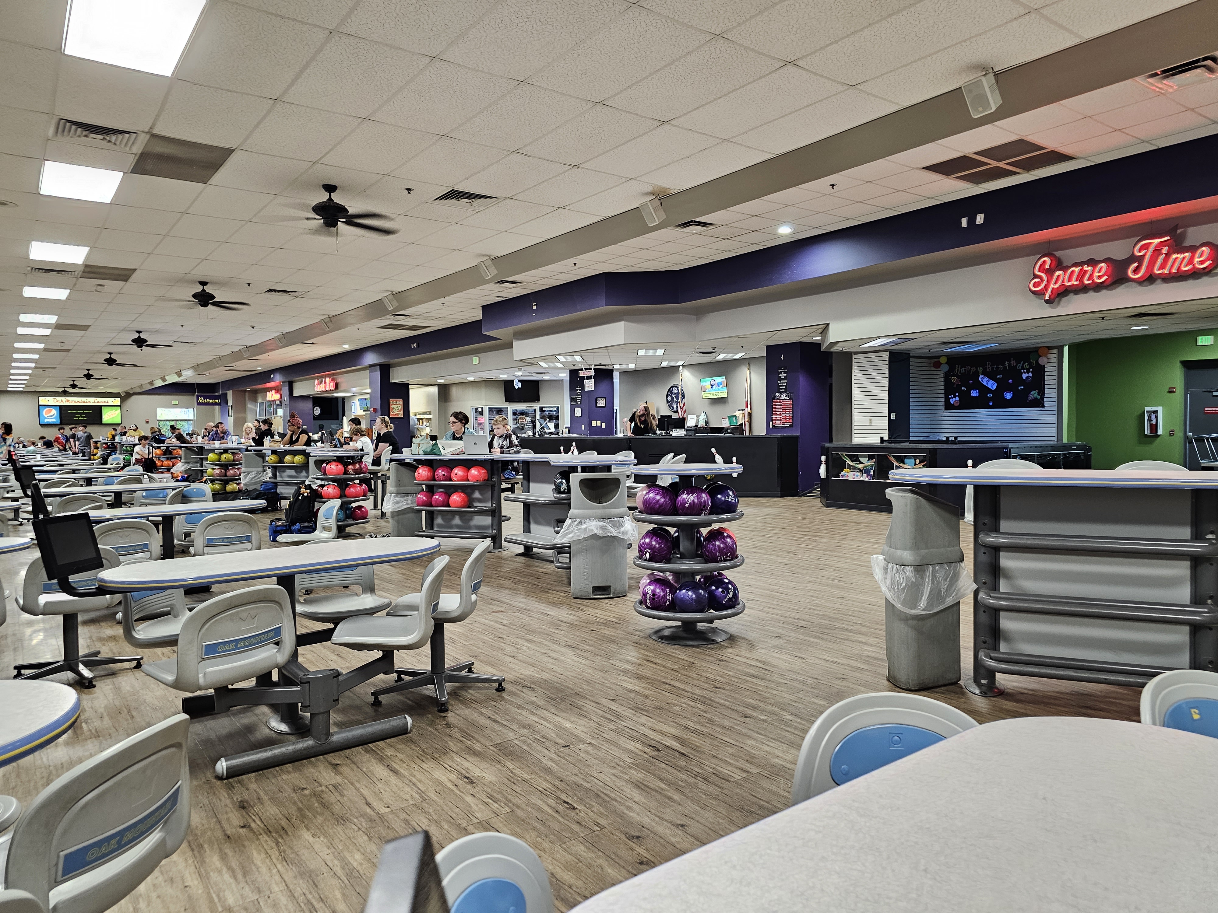 Wide view of a clean bowling center concourse with grey and blue seating, ball racks, and a snack bar area.