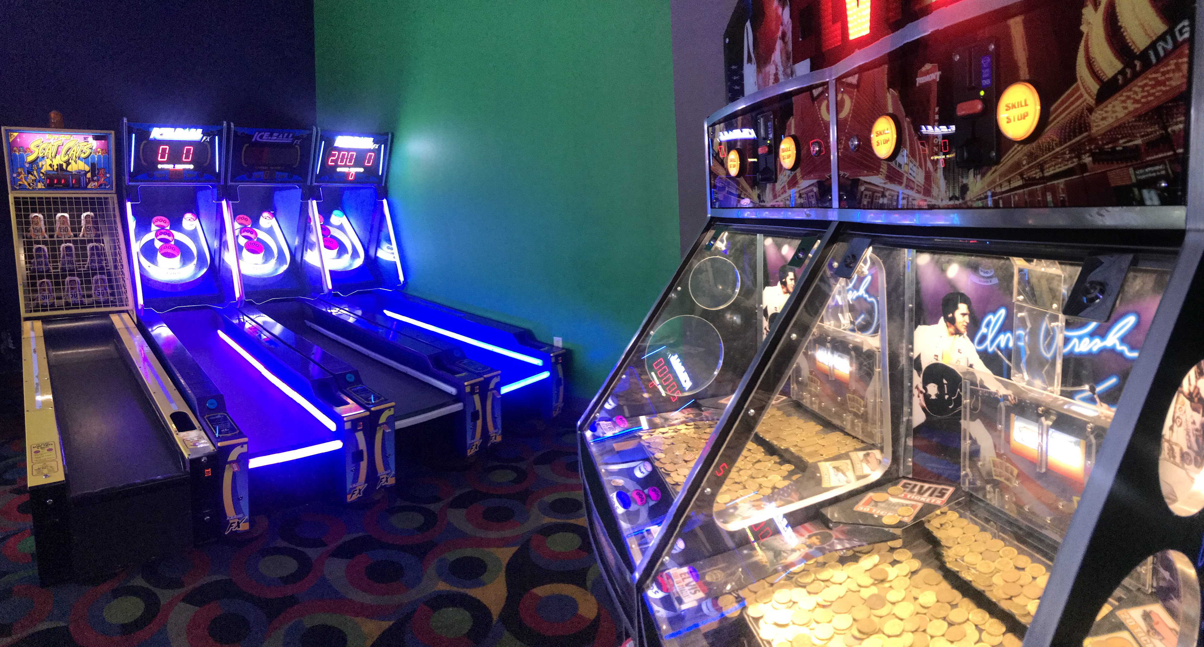 A row of glowing blue and purple Skee-Ball machines next to an Elvis-themed coin pusher game in an arcade.
