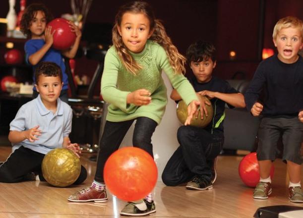 Five young children playing bowling together on a lane with one girl releasing an orange ball.