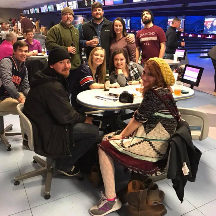 A group of friends smiling and posing around a table at a bowling alley.