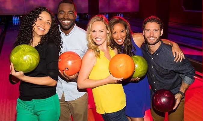 A group of five smiling adults holding bowling balls on a cosmic-lit bowling lane.