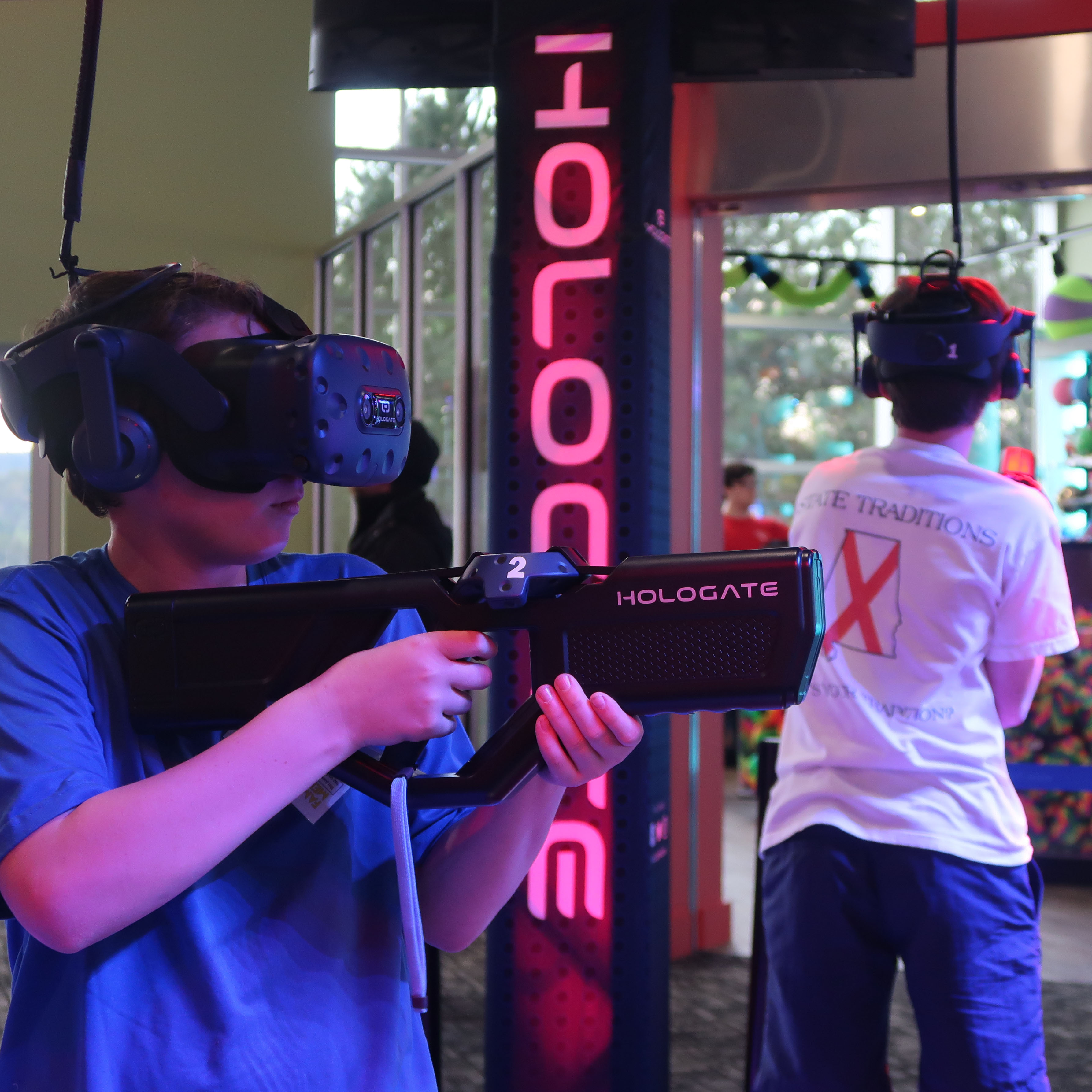 Two people wearing virtual reality headsets and holding haptic controllers at a Hologate VR station.