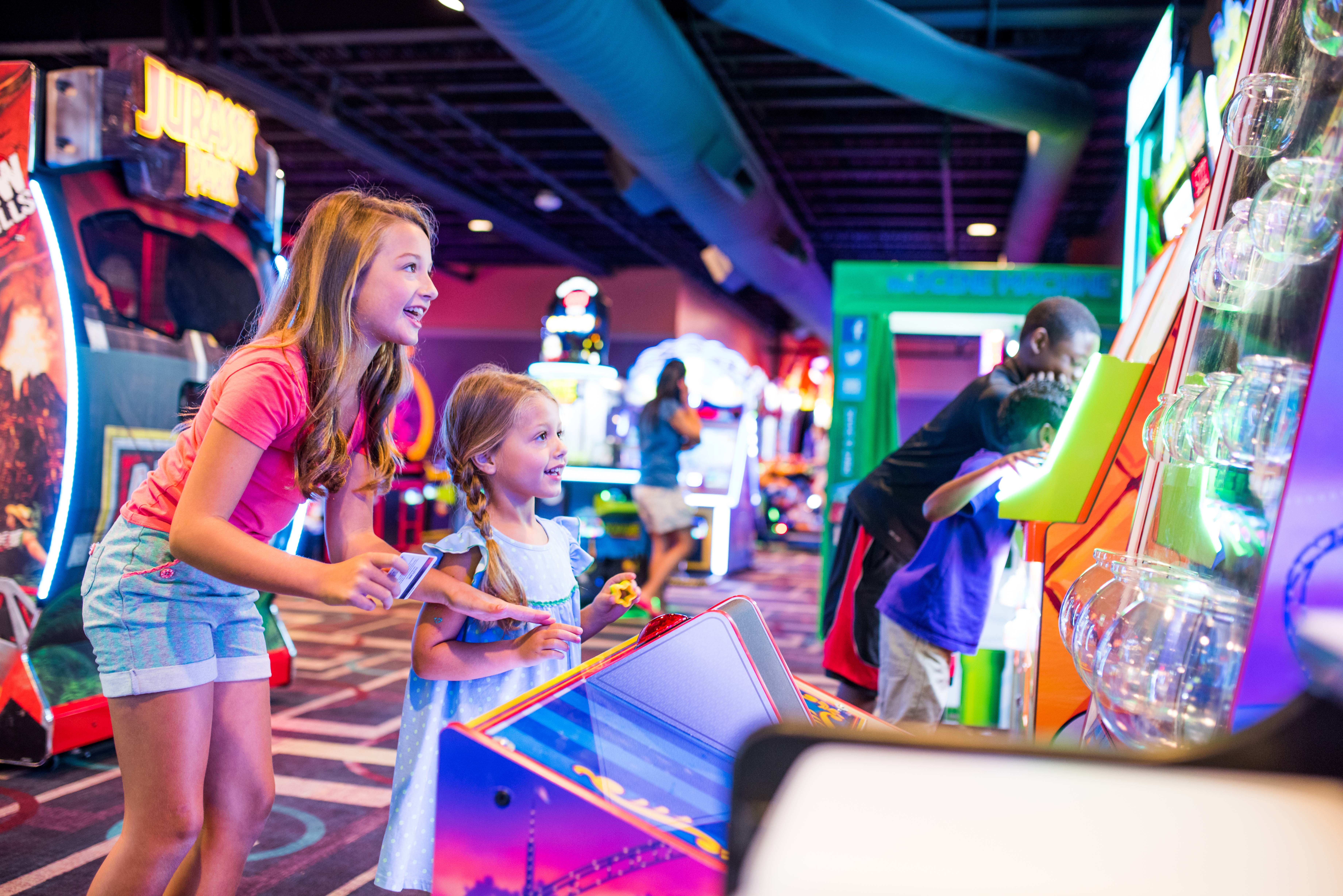 Two young girls smiling and playing a colorful redemption game in a modern arcade.