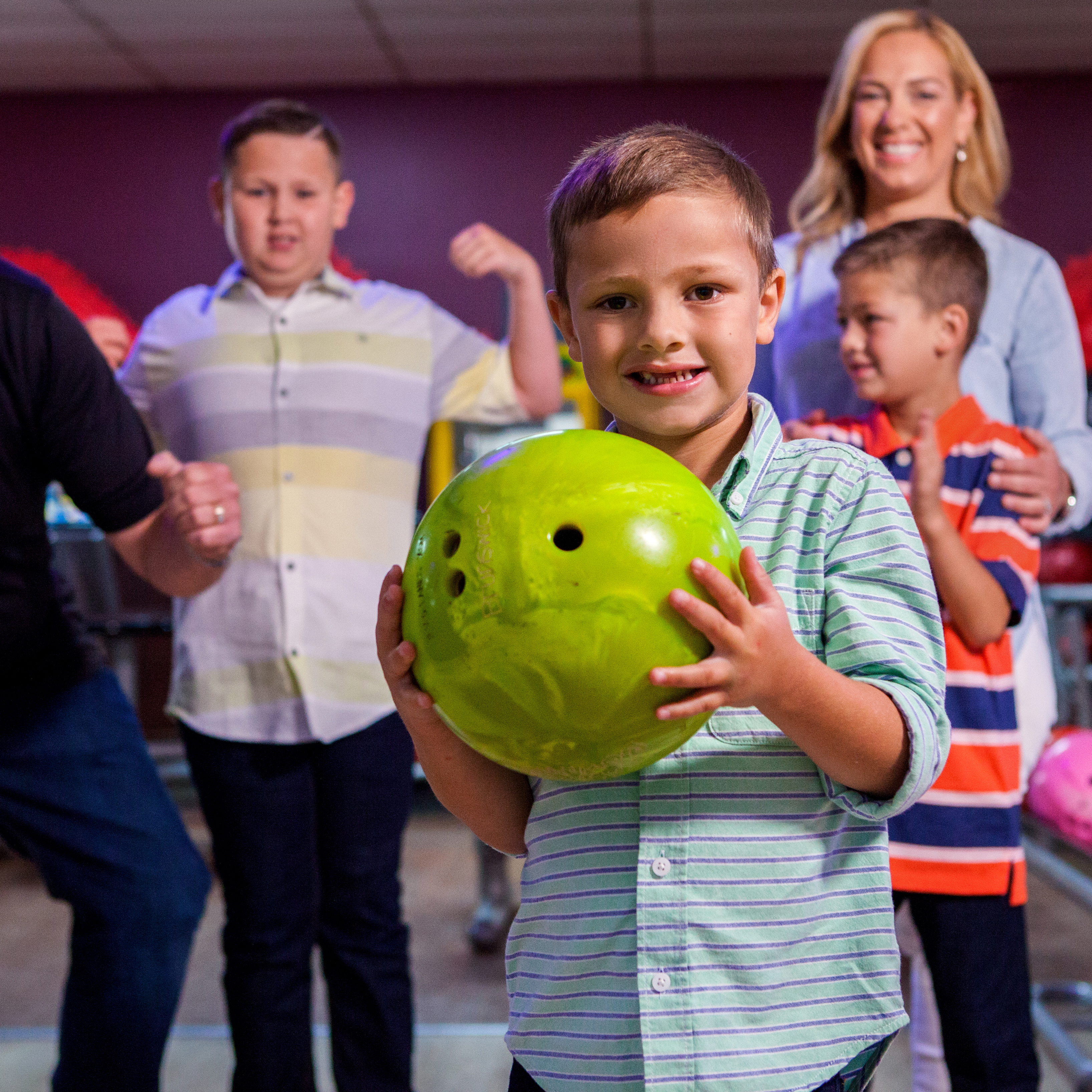 A smiling young boy in a striped shirt holds a bright green bowling ball with friends and family behind him.