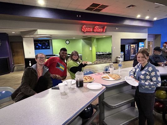 Four people sitting at a counter with pizzas and drinks inside a bowling alley.