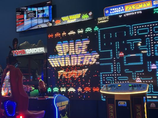Large-screen arcade games including Space Invaders Frenzy and Pac-Man in a dimly lit game room.