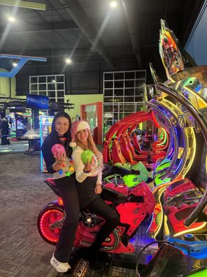 Two women smiling and posing on a red motorcycle racing game in a brightly lit arcade.