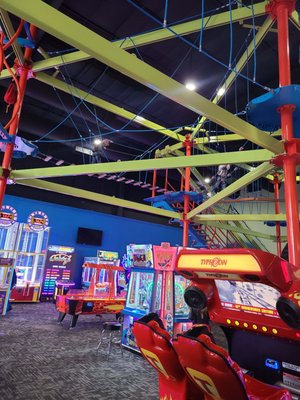 A colorful indoor ropes course structure situated above a variety of arcade games in an entertainment center.