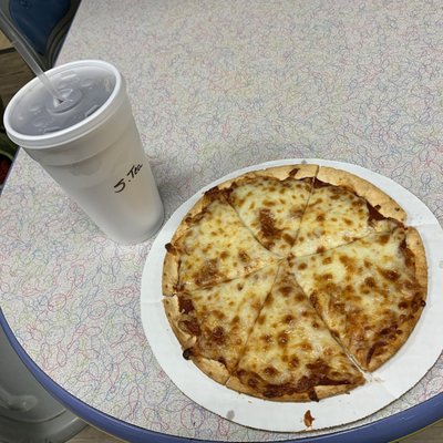 A cheese pizza on a paper plate next to a large foam cup on a patterned table.