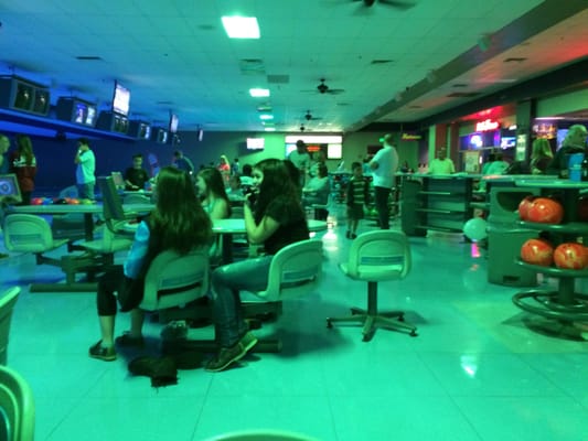 People sitting and standing in a bowling center settee area under bright green and blue cosmic lighting.