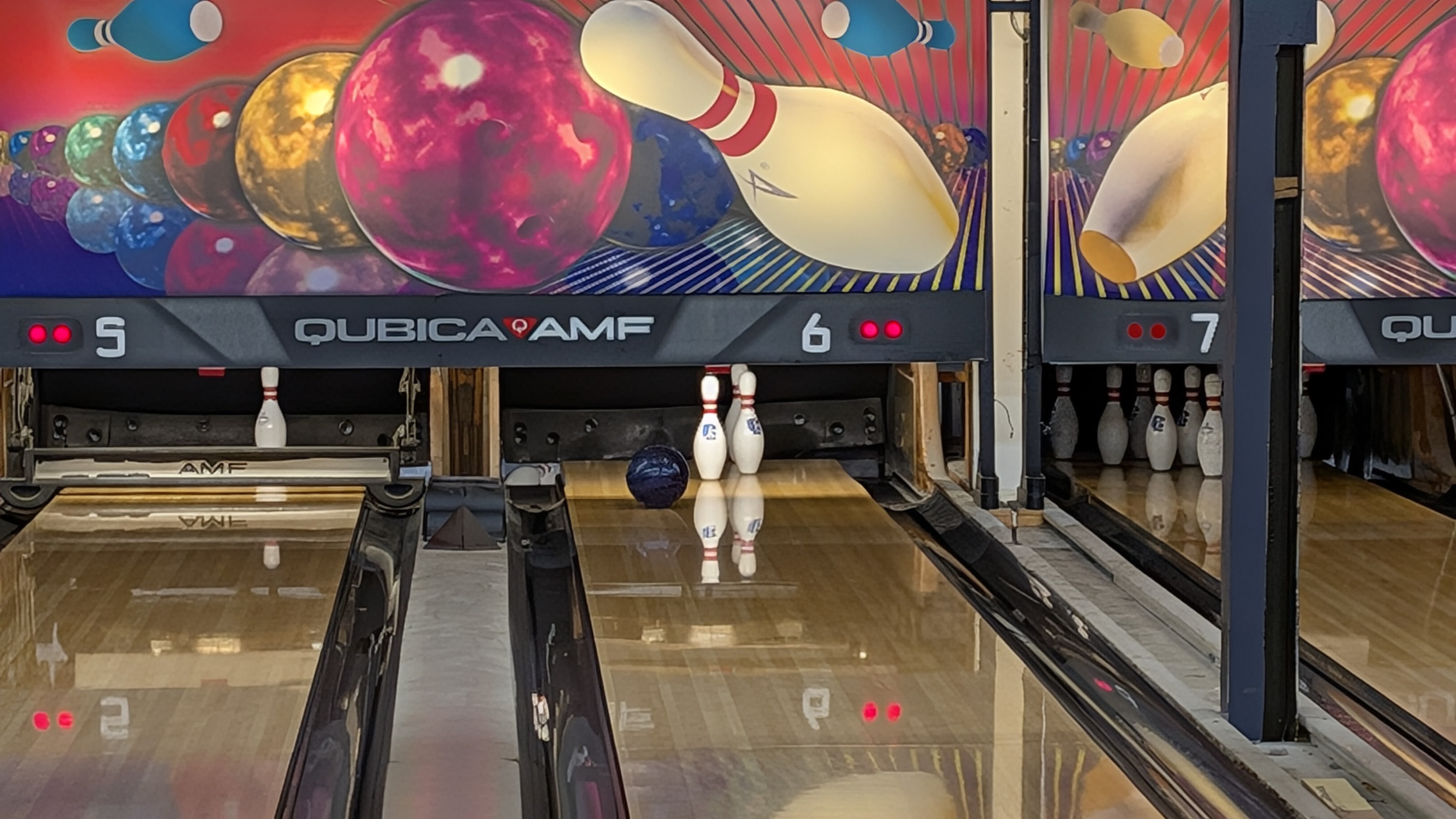 A bowling ball rolls down a polished wooden lane toward two pins under colorful QubicaAMF graphics.