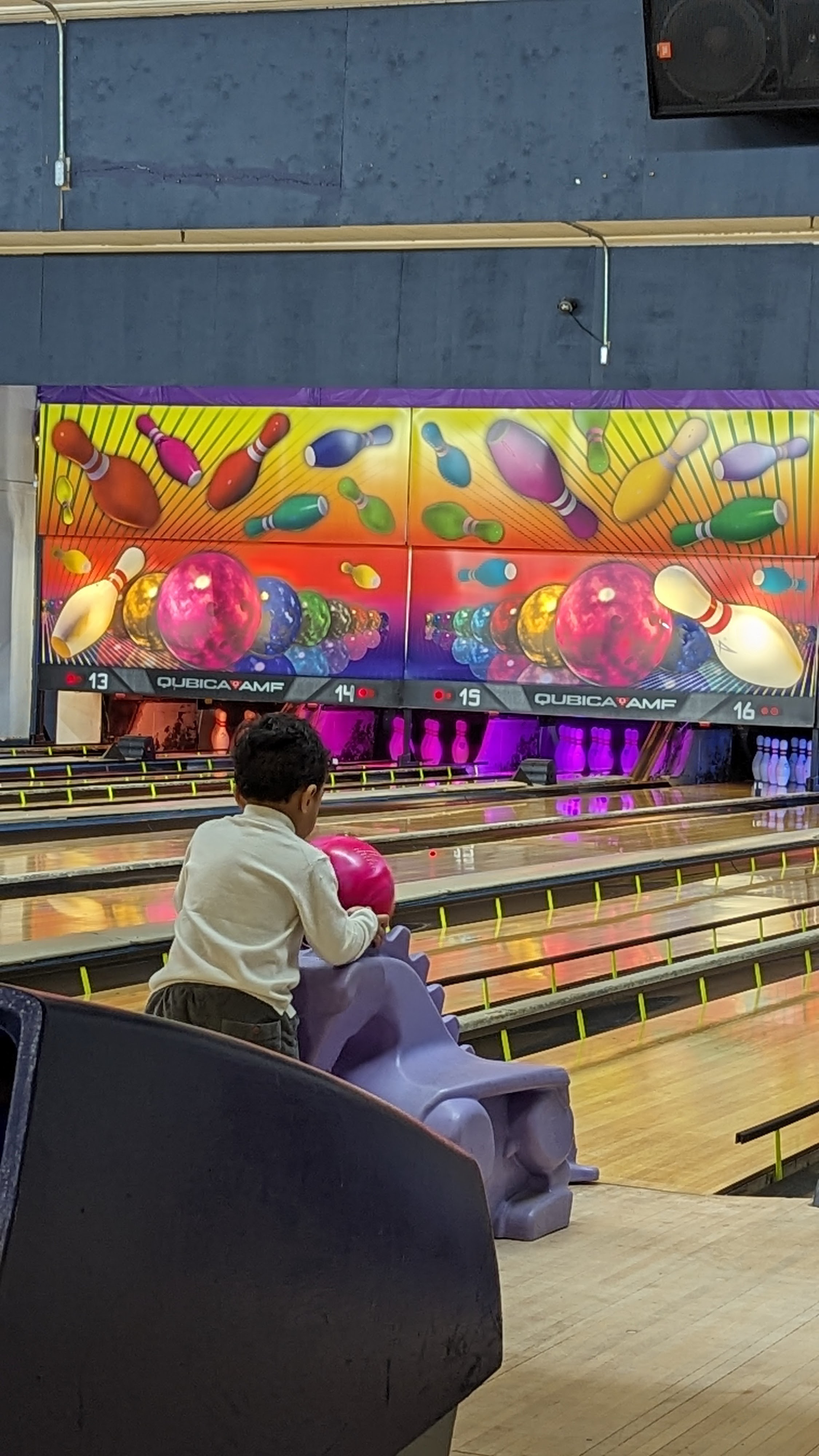 A young child uses a purple dinosaur ramp to bowl a pink ball on a lane.