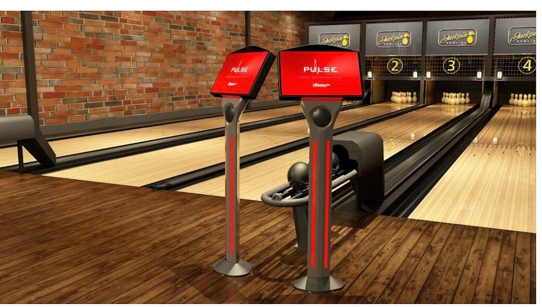 Modern duckpin bowling lanes featuring red touchscreen scoring kiosks and an industrial exposed brick wall.