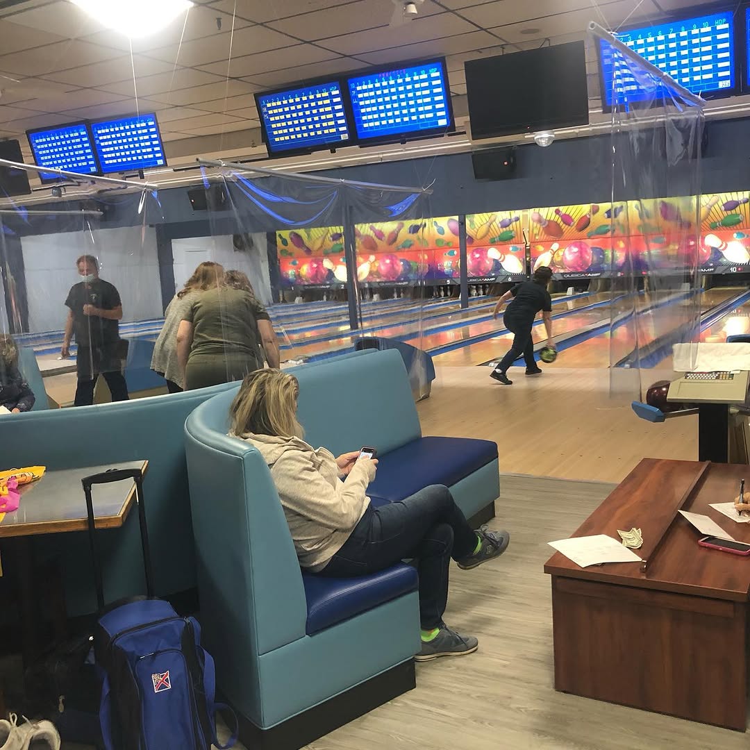 A woman sits in a blue booth while a person bowls on a lane in a brightly lit bowling alley.