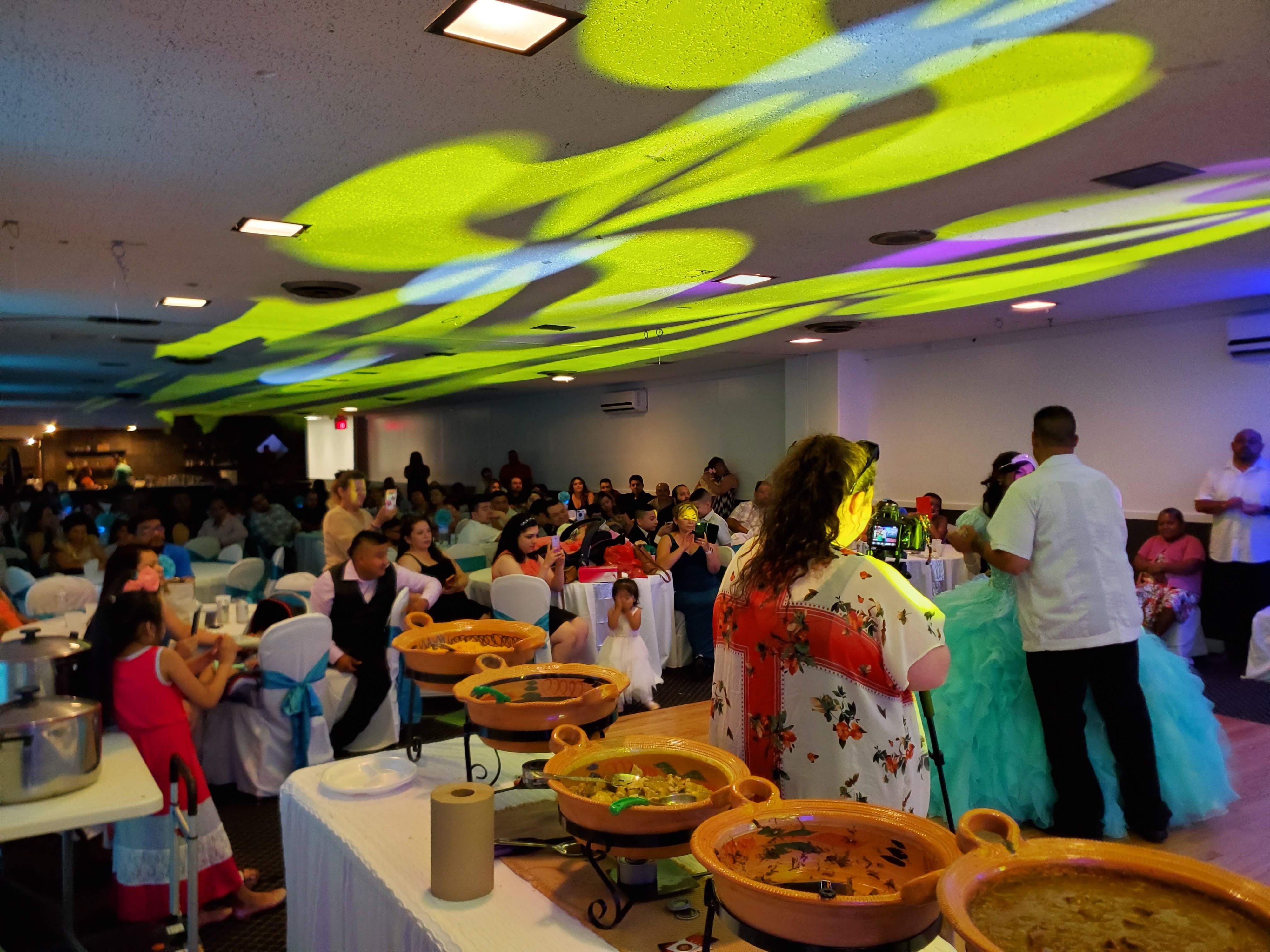 A lively party with a buffet, guests seated at decorated tables, and colorful projected lighting on the ceiling.